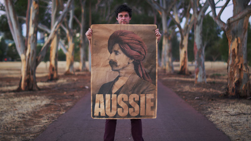 Peter Drew standing on a path in a park holding the 'Aussie' poster