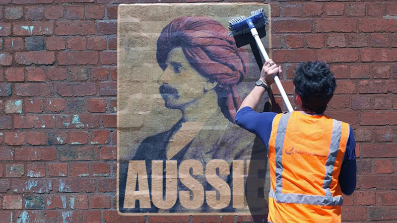 Peter Drew pasting an 'Aussie' poster on a brick wall