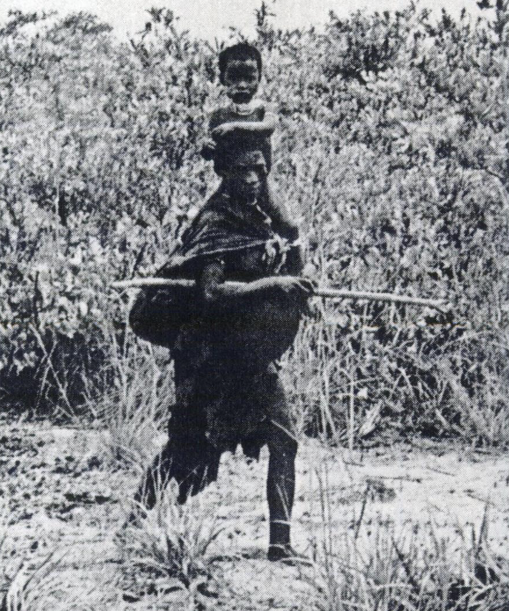 A pregnant woman carrying a shild on her shoulder along with a bag and a digging stick