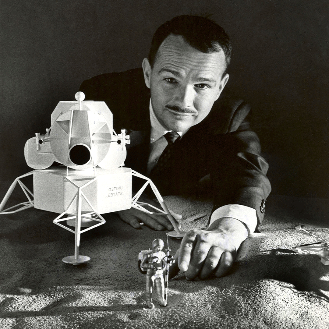 A man crouched behind a table with a model of the moon and a spaceship laid out in front of him
