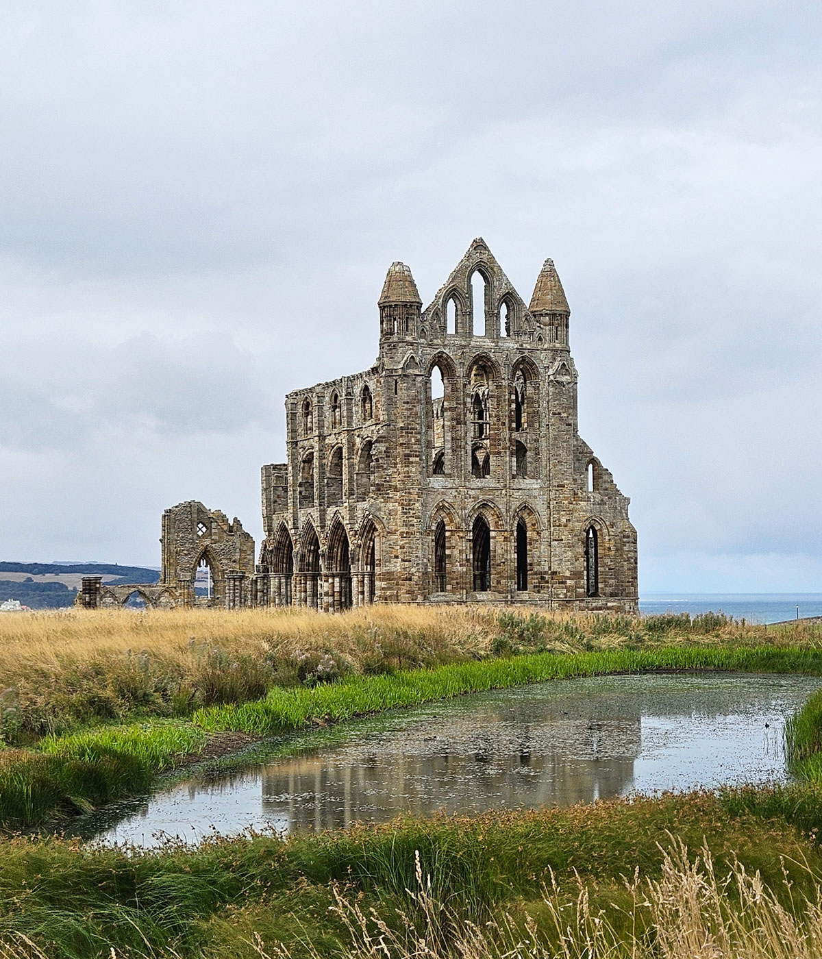 The ruined Whitby Abbey from behind, looking towards the ocean. There is a creek in the foreground, reflecting part of the ruin