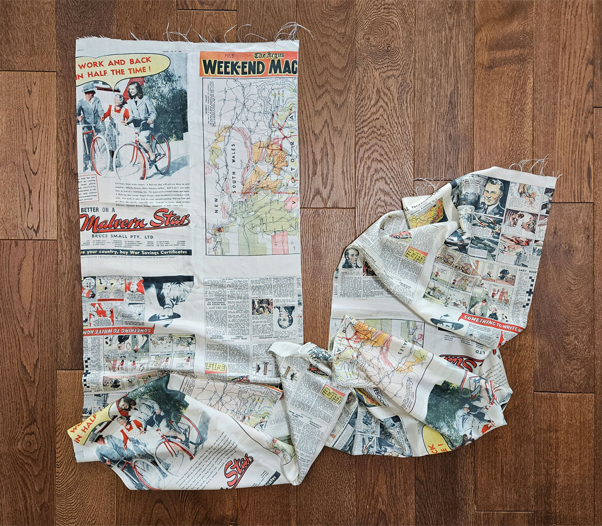 A long piece of silk printed with newspaper pages layed out on a wooden floor