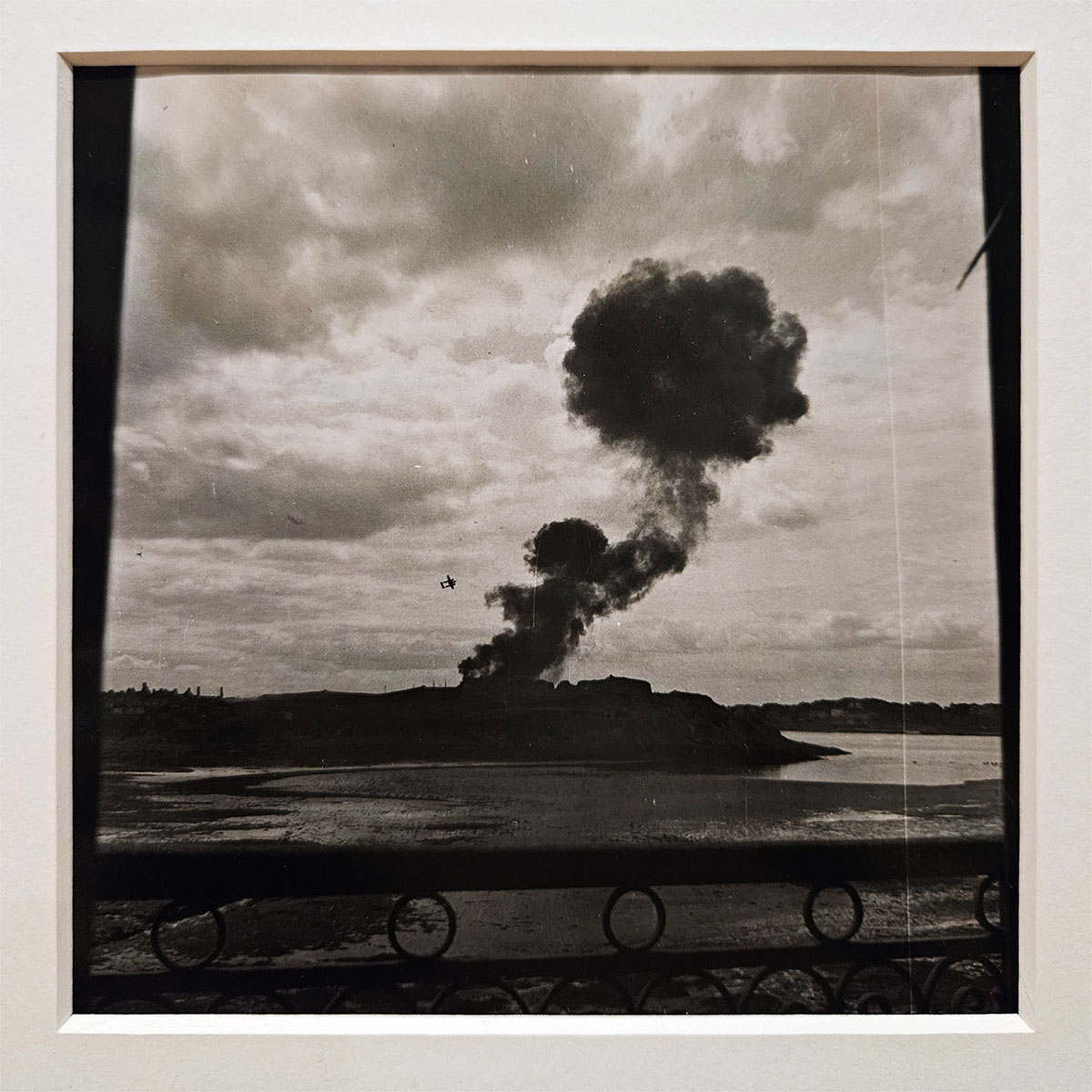 A black and white photograph of a plume of smoke rising from land in the distance, with a plane visible in the sky