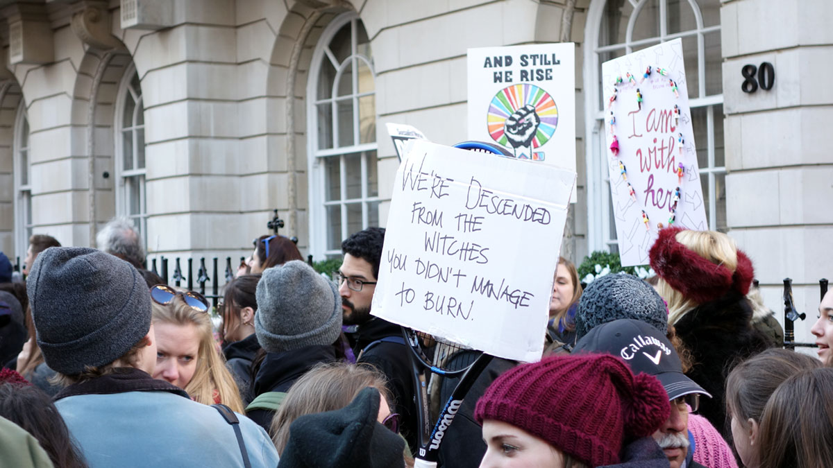 A crowd at a protest march with one person holding a sign that says 'we're descended from the witches you didn't manage to burn'.