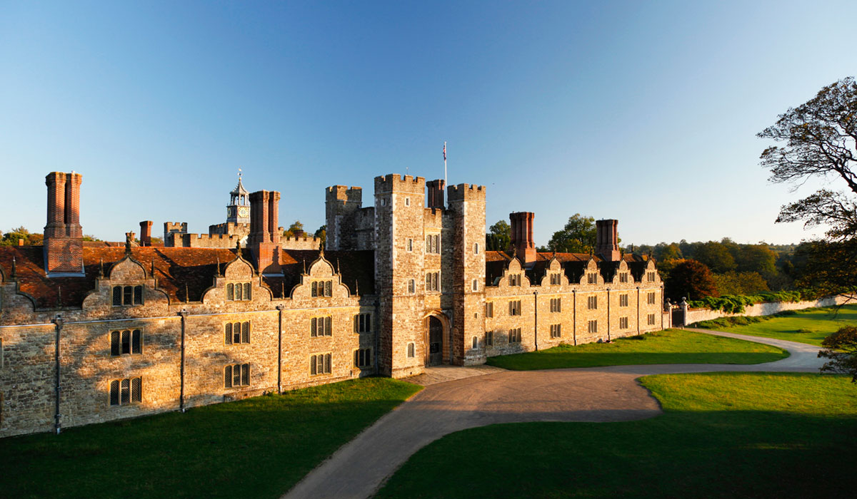 A stately home with a driveway in front of it and trees casting shadows on the brickwork