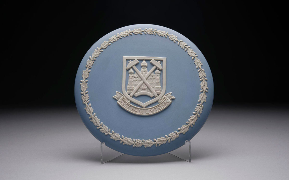 A blue ceramic plate featuring the crest of a football club in white in the centre and a white floral border around the edge