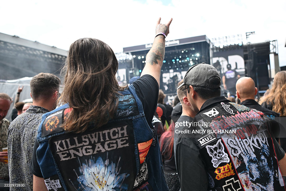 Two men in the foreground of a crowd watching a musical performance on a distant stage. The men have their backs to the viewer and are wearing sleeveless denim jackets decorated with embroidered patches.